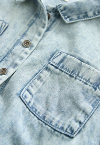 Ljusblå denimskjorta med klassisk krage, framficka, metallknappar och subtilt blekt mönster.