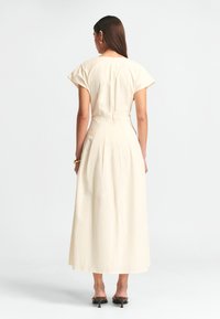 Pitkä kremmäinen mekko, jossa on hihattomat hihat, istuva vyötärö ja valuva hame. Varustettu piilossa olevalla takavetoketjulla ja pehmeällä, sileällä kankaalla.