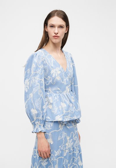 Kvinde med langt brunt hår iført lyseblåt bluse- og nederdelssæt med hvidt blomstermotiv og muslingeskalsbroderi, stående mod hvid baggrund.