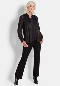 Zwarte doorzichtige button-up shirt over een zwart top, gecombineerd met zwarte op maat gemaakte broeken en enkellaarsjes, wat een gelaagde, monochromatische look accentueert.