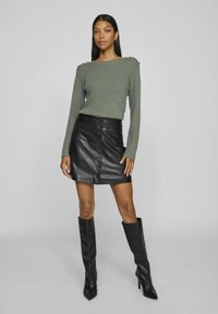 Khaki geribbelde sweater met schouderknopen, gecombineerd met een zwarte leren rok met knopen aan de voorkant en kniehoge zwarte stiletto laarzen.