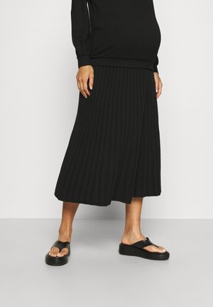 Gravid person iført sort plisseret midilang nederdel, sort langærmet top og sorte platformsandaler mod en ensfarvet baggrund.