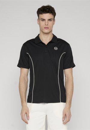 Ung mand står vendt mod fronten iført en sort poloshirt med hvide detaljer og hvide bukser mod en ensfarvet baggrund.