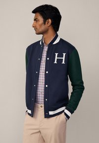 Marine varsityjack met groene mouwen, witte gestreepte kraag en een opvallend 'H' logo. Draagt over een geruit shirt en beige broek.