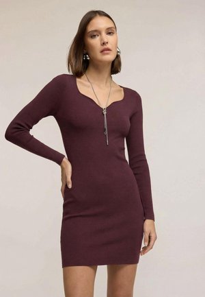 Robe ajustée bordeaux à manches longues avec un décolleté en cœur. Confectionnée en tissu maille côtelée, stylisée avec un collier pendentif argenté. Texture lisse.