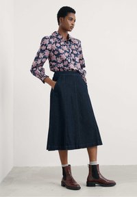 Donna che indossa una blusa floreale rosa infilata in una gonna midi di jeans scuri a vita alta, con stivali alla caviglia marroni e calzini grigi, in piedi.