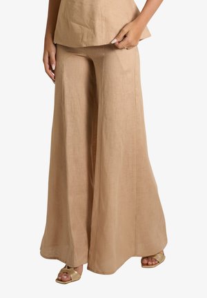Pantaloni beige a gamba larga abbinati a un top coordinato, completati con sandali dorati open-toe e un anello alla mano sinistra.