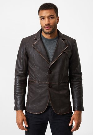 Braune Lederblazer mit taillierter Passform, featuring einen Reverskragen, zwei Knöpfe und kontrastierender Naht. Über einem grauen Pullover getragen.