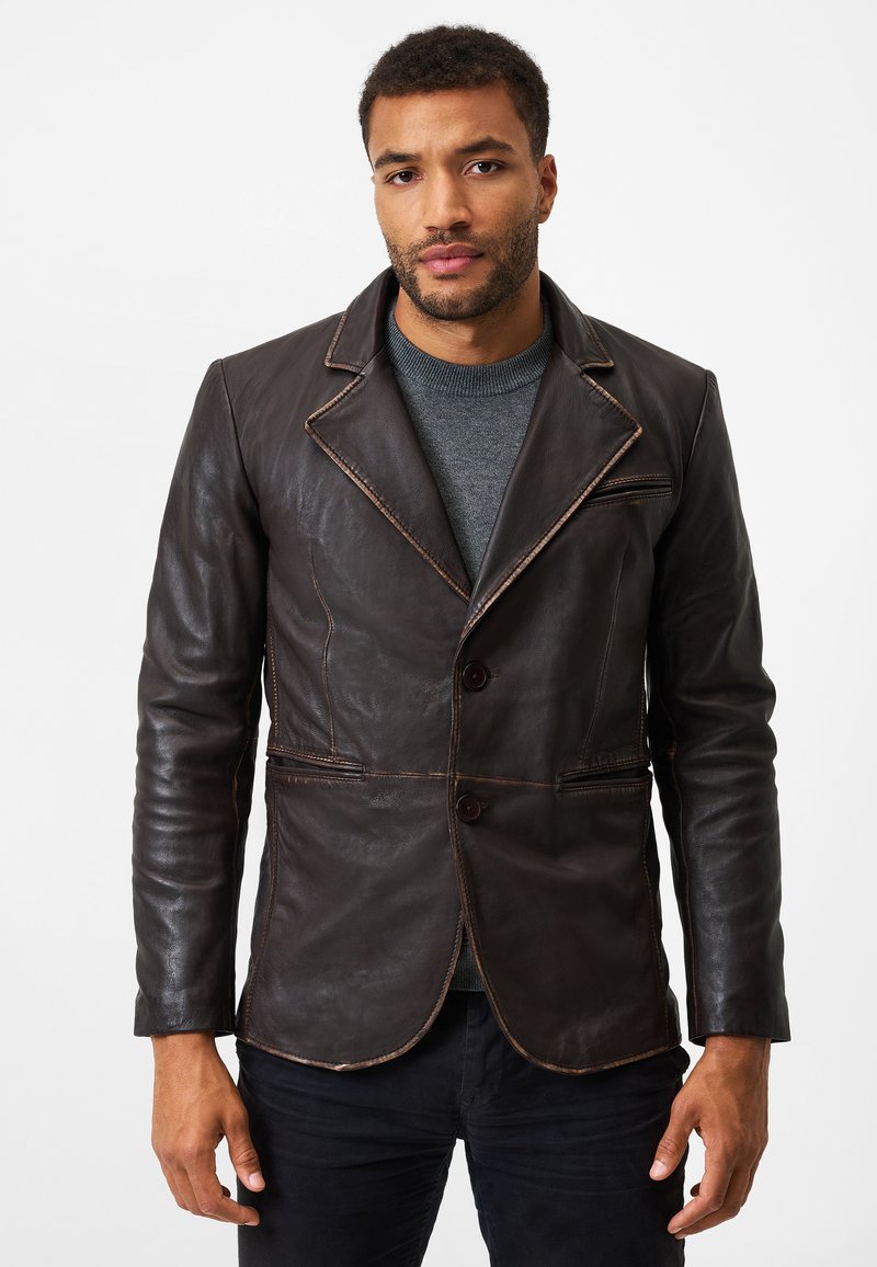 Braune Lederblazer mit taillierter Passform, featuring einen Reverskragen, zwei Knöpfe und kontrastierender Naht. Über einem grauen Pullover getragen.