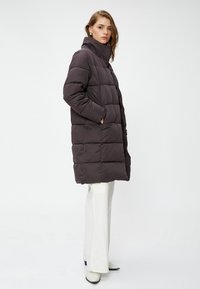 Mujer vestida con un abrigo acolchado largo de color marrón oscuro y cuello alto, pantalones blancos de pierna ancha y botas blancas de punta, de pie de perfil.