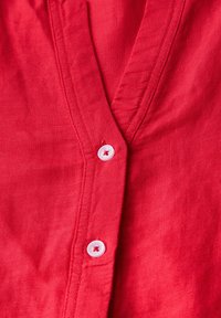 Camicia di lino rossa con scollo a V, cuciture visibili e un bottone bianco al colletto. Texture liscia con un aspetto leggero.
