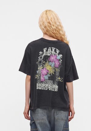 Personne aux longs cheveux blonds porte un t-shirt noir oversize avec un motif floral et un texte dans le dos, associé à un jean cargo bleu.