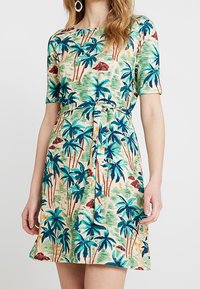 Kratké šaty s krátkým rukávem vyrobené z zelené látky s barevným vzorem palmových stromů. Obsahují detail vázání v pase a rozšířený lem.