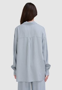 Jasnoszara, długorękawowa koszula o luźnym kroju, zaokrąglonym dole i szczególe w postaci plisowanej pleców. Posiada kołnierzyk i mankiety zapinane na guziki.