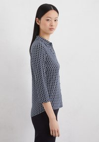 Marineblaue Bluse mit weißem geometrischen Muster, Kragen und Dreiviertelärmeln. Glattes Material, leicht taillierte Form und Knopfleiste.
