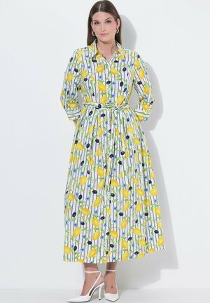 Kvinde iført en lang hvid kjole med gult citron-, blåt blomstermønster og oliventræsmotiv, trekvartærmer og hvide højhælede sko.