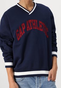 Granatowa bluza z dekoltem w serek, czerwony napis „GAP ATHLETIC”, biały kołnierz i mankiety w paski, miękki materiał, luźny krój i ściągacz na dole.