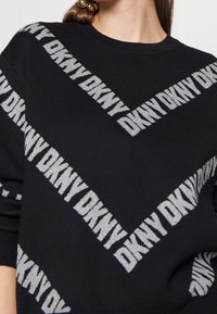 Schwarzer Pullover mit diagonalen DKNY-Logo-Mustern in Silber. Verfügt über einen Rundhalsausschnitt und gerippte Bündchen, die sowohl Textur als auch Design betonen.