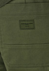 Parche de tela verde oliva con el texto "WORKWEAR STUDIO" en negro, cosido sobre un fondo texturizado con costuras visibles.