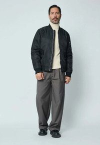 Veste en cuir noire avec un col en tricot côtelé, pull col roulé beige et pantalon ample gris, accompagnés de chaussures noires. Fond uni.