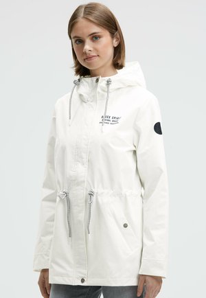 Young woman wearing a white hooded jacket with drawstrings, side pockets, and logo patches on chest and sleeve, standing against a plain background.