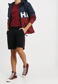 Námořnická a jemně červená zipová bunda s velkým bílým logem, červené tričko, černé šortky s červeným bočním pruhem a burgundské tenisky.