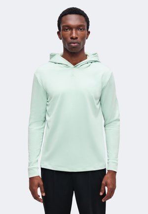 Mann trägt hellgrünes Kapuzenshirt mit langen Ärmeln und schwarze Hose, steht vor einem einfarbigen hellgrauen Hintergrund.
