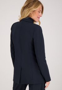 Marineblauwe op maat gemaakte blazer met gestructureerde vorm, lange mouwen en een rugventilatie-detail. Zachte stoftextuur met een gladde afwerking.