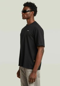 Uomo che indossa una t-shirt nera oversize di G-Star, jeans grigi e occhiali da sole neri rettangolari, in piedi contro uno sfondo grigio chiaro, profilo laterale.