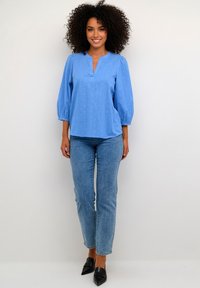 Svetlomodrá blúzka s výstrihom do V, nafúknutými rukávmi a texturovaným vzorom; kombinovaná so svetlomodrými džínsami a čiernymi topánkami s hrotom.