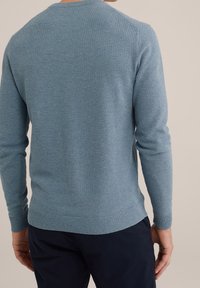 Man som bär en texturerad ljusblå långärmad tröja och mörkblå byxor, sedd bakifrån mot en enfärgad bakgrund.