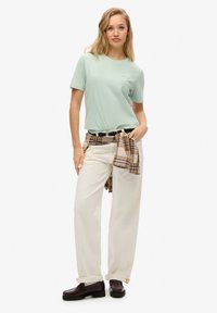 Camiseta de manga corta verde claro, pantalones crema de corte relajado y zapatos oscuros. Una camisa de cuadros atada a la cintura añade un detalle en capas.