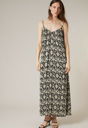 Femme portant une robe maxi noire et blanche à motifs géométriques sans manches avec de fines bretelles et des sandales noires à talons, debout devant un fond uni.