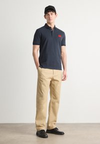 Marineblå polo skjorte med korte ermer og en rød logo, kombinert med beige khaki bukser og svarte sko. Glatt stoff og skreddersydd passform.