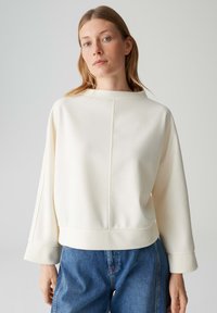 Roomkleurige, boxy sweatshirts met een hoge halslijn, voorzien van een naaddetail en brede mouwen, gecombineerd met blauw losvallende jeans.
