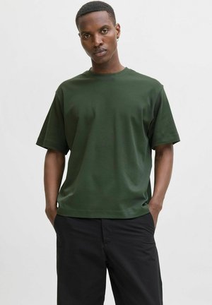 T-shirt vert foncé à manches courtes en mélange de coton, avec un col rond et une coupe décontractée. Porté avec un pantalon noir.