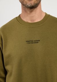 Oliwkowy sweter z okrągłym dekoltem, z małym czarnym nadrukowanym logo na piersi. Gładka tekstura materiału.
