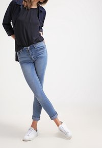 Jeans de un azul claro con un corte slim, dobladillos enrollados y diseño de cinco bolsillos, combinados con una blusa negra y zapatillas blancas.