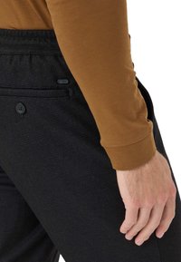 Zwarte textuurbroek met knoopdetails, gecombineerd met een bruine langemouwenshirt. Zichtbare hand rustend op de heup, die de stofcontrast laat zien.