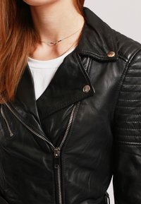 Giacca da biker in pelle nera con dettagli trapuntati sulle spalle, hardware argentato e chiusura con zip asimmetrica. Indossata sopra una camicia bianca.