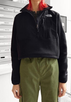 Osoba o rudych włosach, ubrana w przyciętą czarną bluzę polarową North Face oraz oliwkowe spodnie cargo, stoi w pomieszczeniu obok skrzynek na listy.