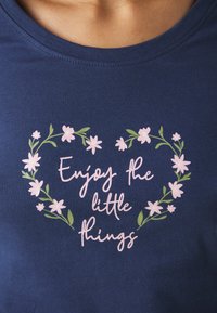 Mörkblå t-shirt med rund halsringning; har en rosa blommönster som omger texten "Njut av de små sakerna" i kursiv stil.