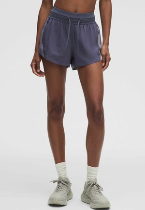 Atleet draagt donkergrijze sportshorts, witte sokken tot halverwege de kuit en lichtgrijze hardloopschoenen en staat op een effen achtergrond.