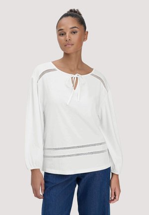 Frau trägt eine weiße Bluse mit langen Ärmeln, einer Krawatte am Ausschnitt und Mesh-Details an Schultern und Saum, kombiniert mit blauen Jeans.