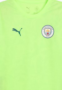 Vihreä urheilupaita lyhyillä hihoilla, jossa on tummansininen Puma-logo ja pyöreä Manchester City -embleemi edessä.