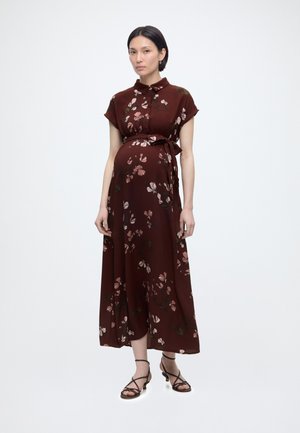 Έγκυος γυναίκα με καφέ midi φόρεμα με λουλουδένιο σχέδιο, κοντά μανίκια και δεμένη μέση, σε συνδυασμό με μαύρα σανδάλια με λουράκια, στέκεται μπροστά σε απλό φόντο.