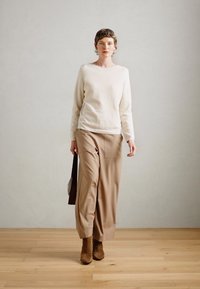 Kremkrāsas gara piedurkne virsū, pārī ar bēšiem platākajiem biksēm un brūnām augstpapēžu zābakiem. Modelis tur tumšu somu; minimālistisks fons ar koka grīdu.
