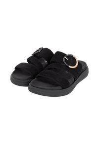 Black suede sandals feature two wide straps and a circular gold and black hardware accent. The sole is flat and made of a textured material.