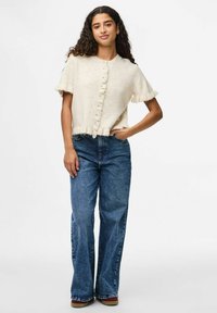 Krémový kardigan s krátkým rukávem a volánovým okrajem, s designem na zapínání na knoflíky, kombinovaný s modrými širokými kalhotami s vysokým pasem.