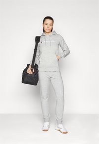Grauer Baumwoll-Hoodie mit Fronttasche, mit "Champion"-Logo. Kombiniert mit passenden Jogginghosen und weißen Sportschuhen. Schwarze Sporttasche.